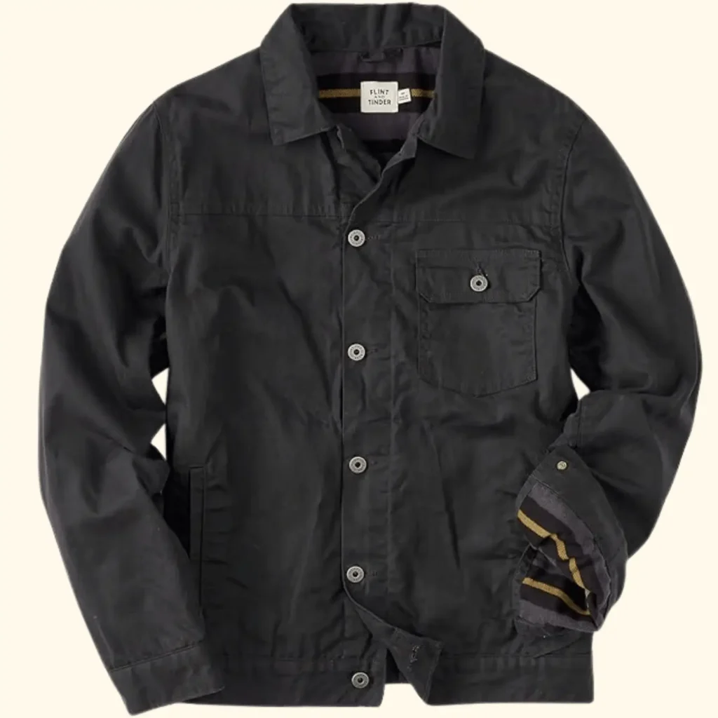 A lifestyle shot of the Flint and Tinder Flannel-Lined Waxed Trucker Jacket in a rugged Field Tan color. The jacket features a durable waxed sailcloth exterior with a classic button-front design, a single button-flap chest pocket, and a soft, striped flannel lining visible at the collar and interior.