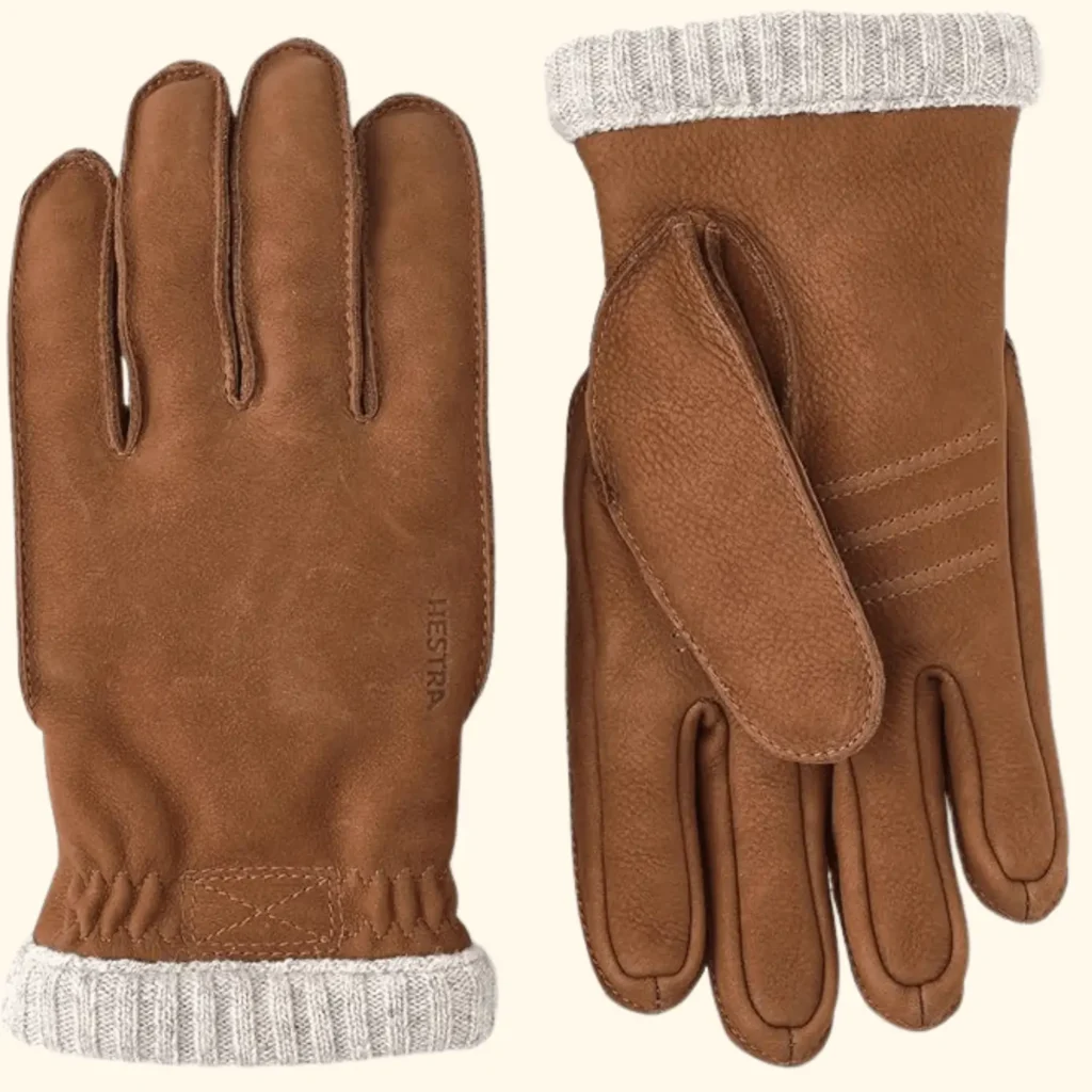 A product shot of the Hestra Utsjo gloves in a warm tan elk leather. The image displays both the front and back of the gloves, highlighting the rugged texture of the Nordic leather, decorative triple-line stitching on the back, and a soft, grey wool-ribbed cuff designed for insulation.