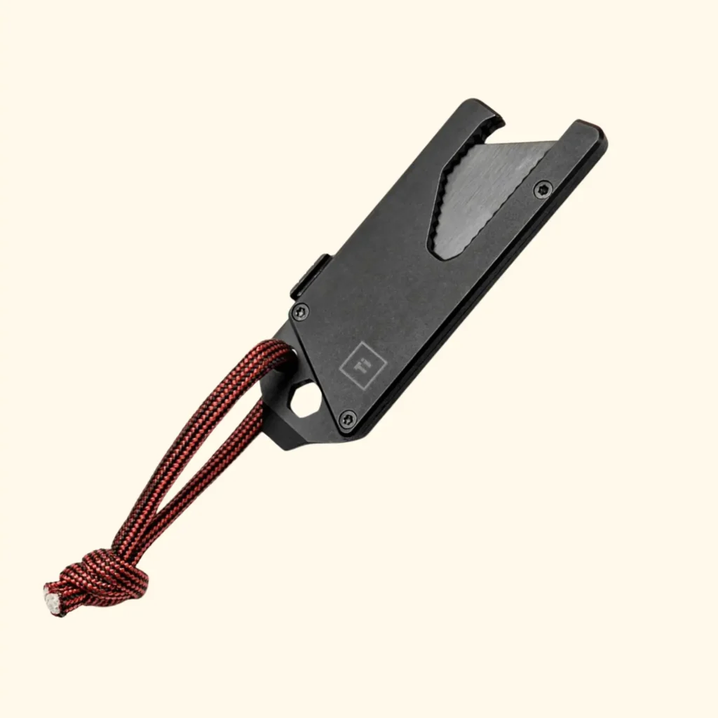 A product shot of the TPT Slide V2 Titanium Utility Tool in a matte black finish. The pocket-sized multi-tool features a slide-out utility blade, a built-in hex wrench at the base, and an attached red and black paracord lanyard for easy retrieval and carry.
