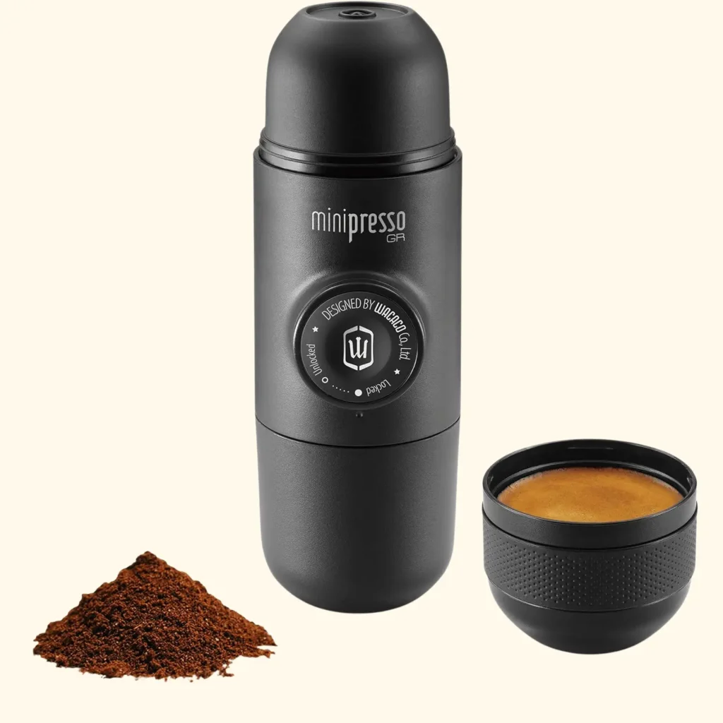 A product shot of the Wacaco Minipresso GR, a compact, pill-shaped portable espresso machine in matte black. The device features a central semi-automatic piston that is unlocked to manually pump and extract coffee, and the removable cap on one end doubles as a built-in espresso cup.