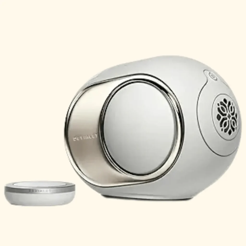 A product shot of the Devialet Phantom I wireless speaker in white, featuring its signature spherical, aerodynamic design and rose gold-plated side panels. Beside the speaker is its dedicated circular remote control, which features a smooth rotary dial for precise volume and playback management.